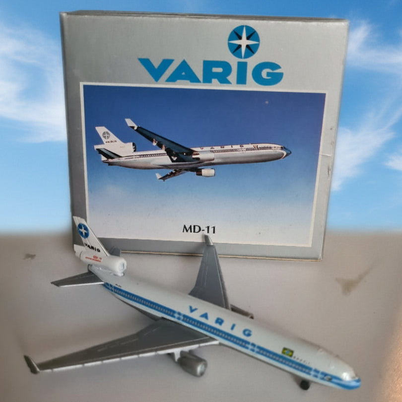 A Varig MD-11 model airplane displayed next to its original packaging box on a shelf. The model is white with blue stripes and tail markings, while the box features a blue and white printed design with the Varig logo.