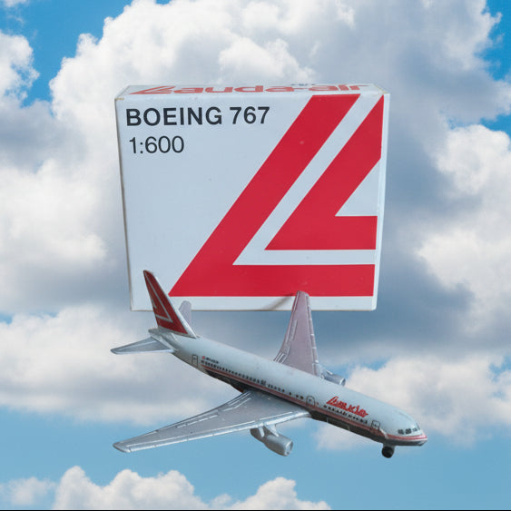 A small white Boeing 767 model airplane beside its branded box, photographed on a plain tan surface. The box shows red branding and text reading Boeing 767 1:600.