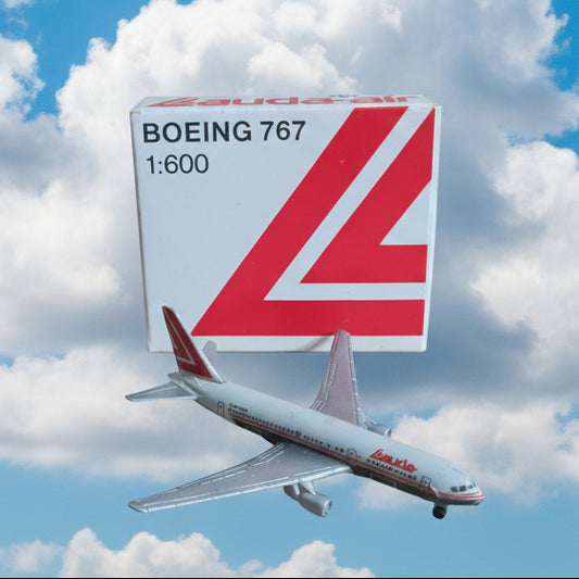 A small white Boeing 767 model airplane beside its branded box, photographed on a plain tan surface. The box shows red branding and text reading Boeing 767 1:600.