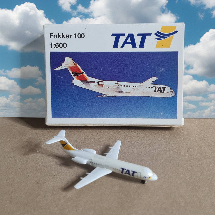 A small white TAT Fokker 100 model airplane placed beside its retail box on a brown cardboard background. The box shows the TAT logo and an illustration of the airplane, with text reading Fokker 100 and 1:600.