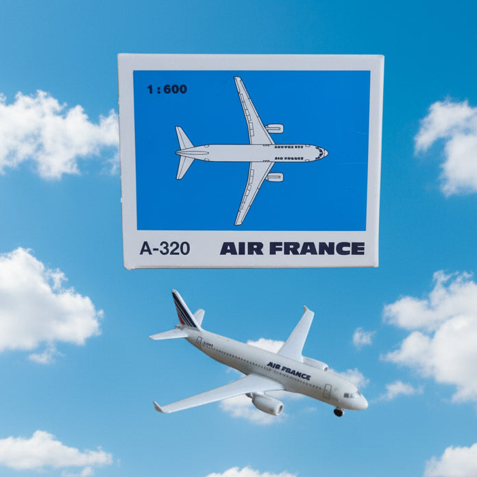 A small white model airplane placed in front of its packaging box. The box has a blue label with a white airplane illustration and the text '1:600 A-320 AIR FRANCE'.