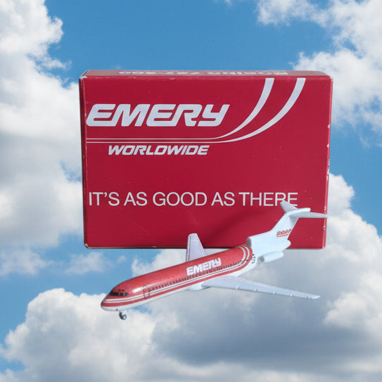 A small model airplane with a red and white fuselage and white wings sits in front of its red packaging box, which displays the brand name 'EMERY WORLDWIDE' and the slogan 'IT'S AS GOOD AS THERE'. The photo is taken on a plain brown cardboard surface.